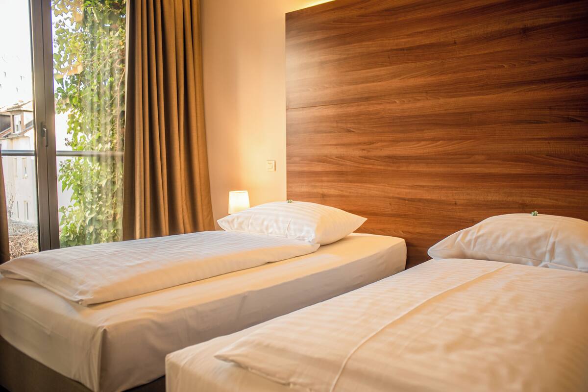A hotel room with two beds, a wooden headboard, a window with curtains, and a view of greenery outside.