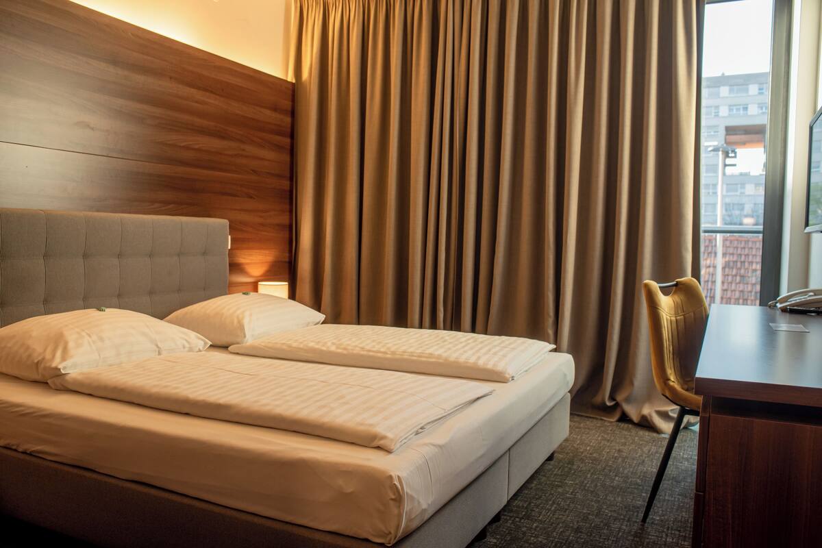 A hotel room with a double bed, a wooden headboard, a desk with a chair, and a view of buildings through the window.