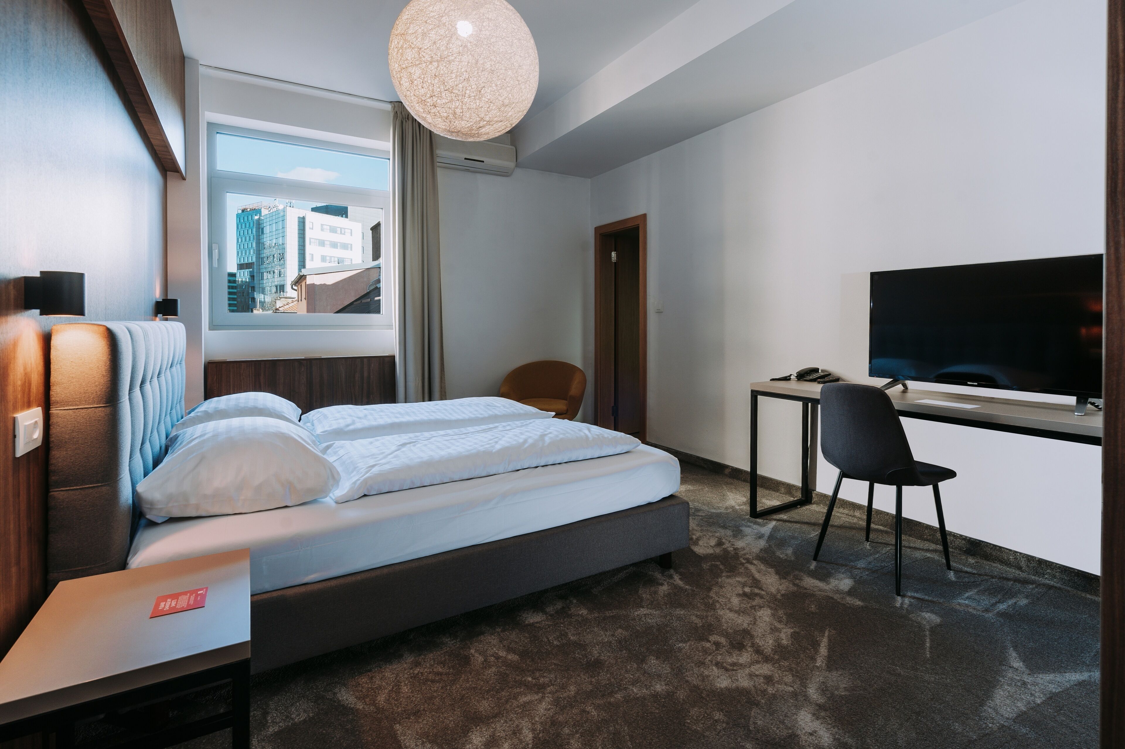 A modern hotel room with a bed, a desk, a chair, a TV, and a window with a city view.