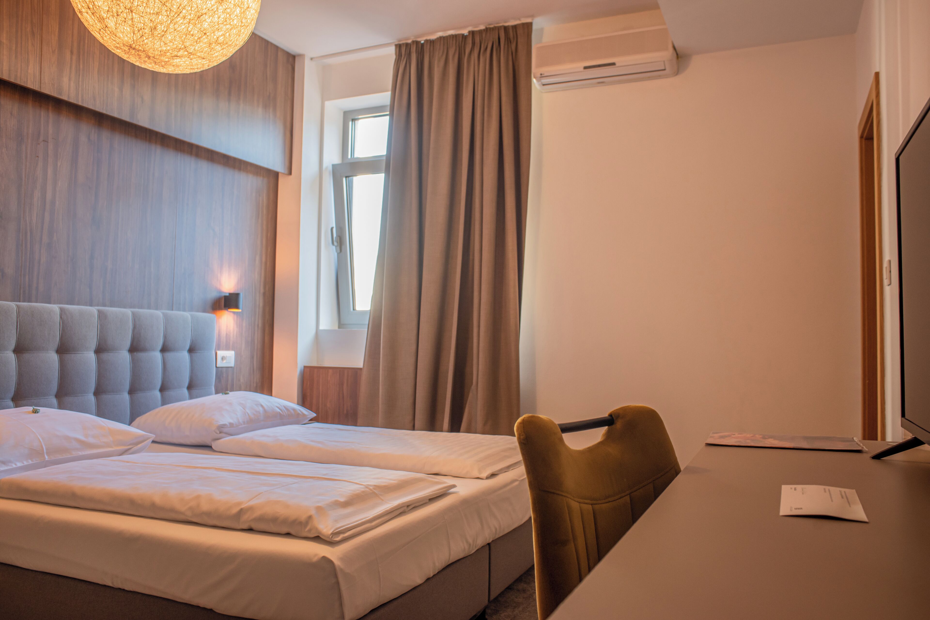 A hotel room with a large bed, a desk, a chair, a lamp, and an air conditioning unit.