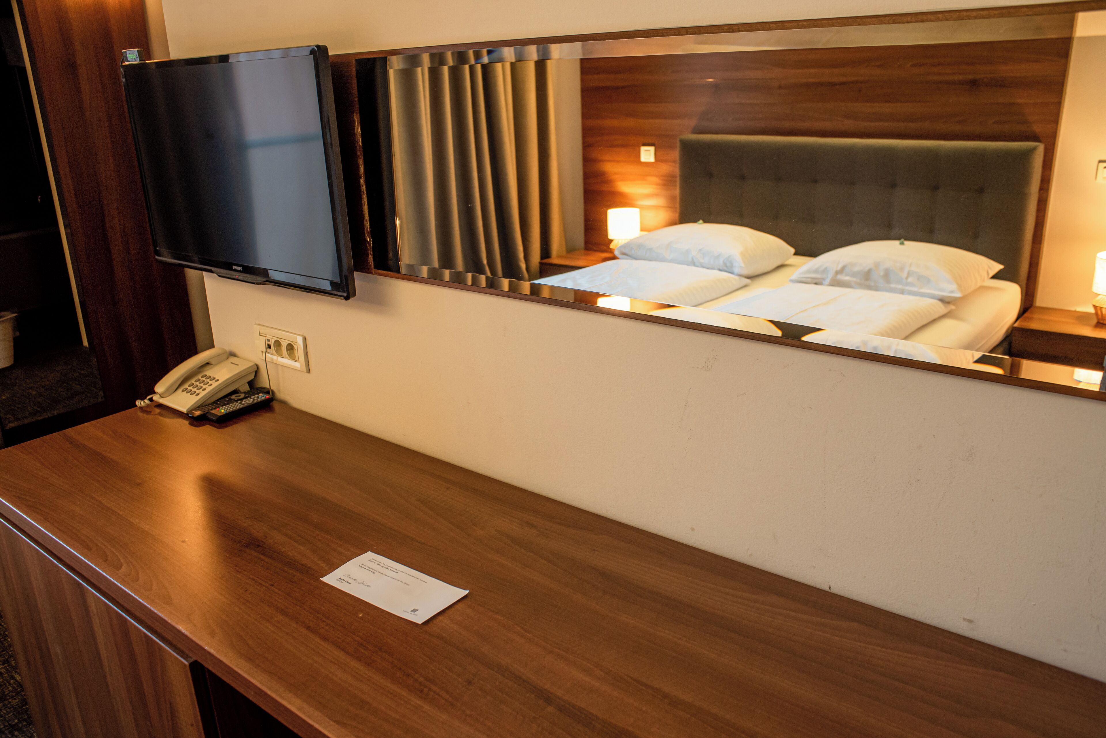 Een hotelkamer met een houten bureau, een flatscreen televisie aan de wand, een telefoon en een bed met witte lakens.