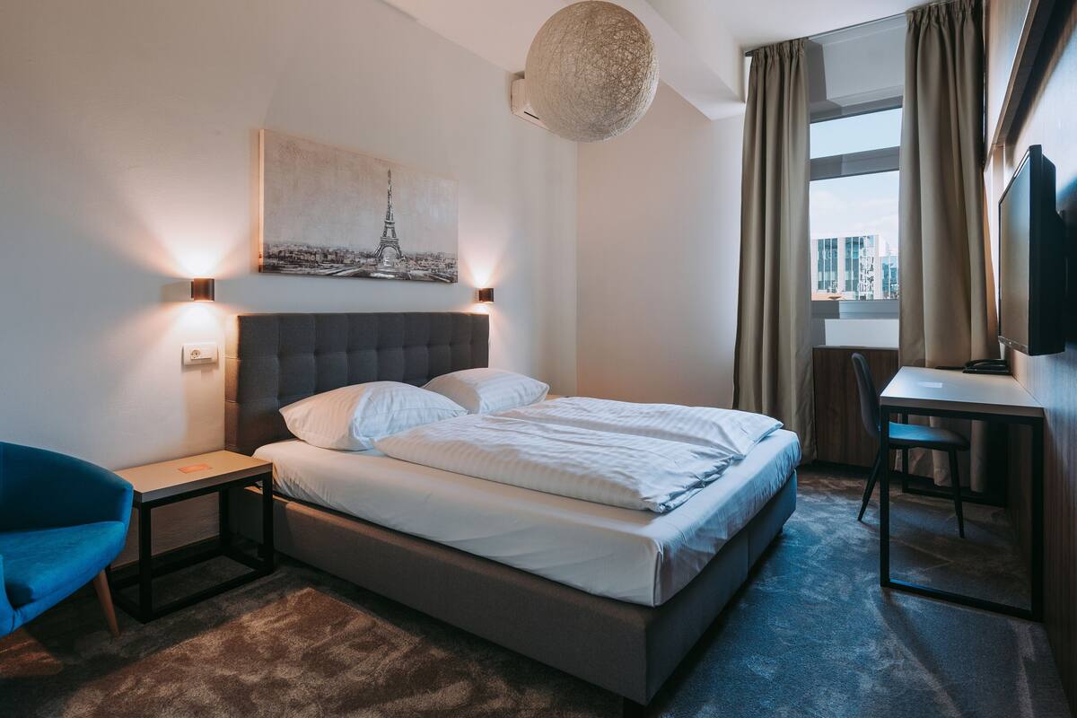A hotel room with a bed, a desk, a chair, a lamp, a painting of the Eiffel Tower, and a view of the city through the window.