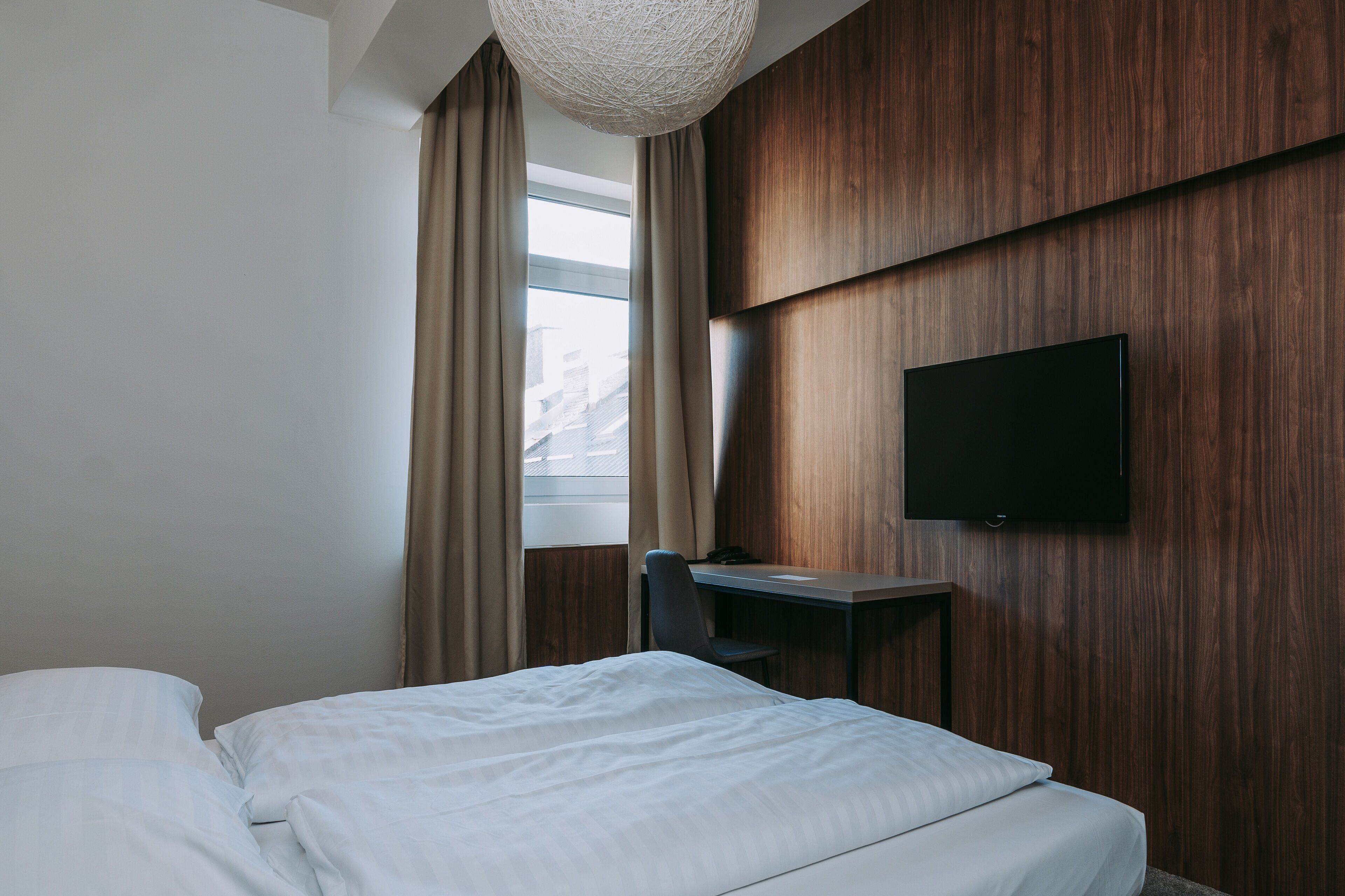 A hotel room with a bed, a desk, a chair, a television mounted on the wall, and a window with curtains.