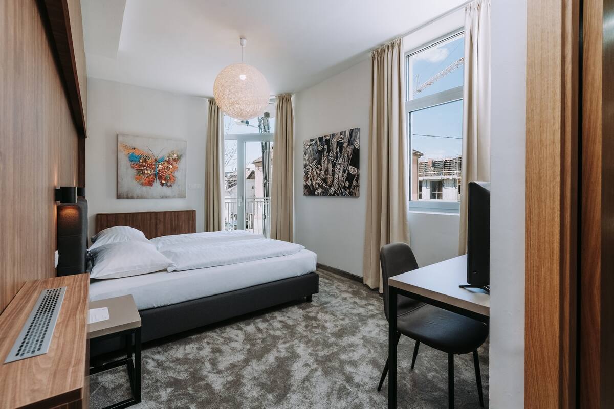 A hotel room with a bed, desk, chair, and large windows.