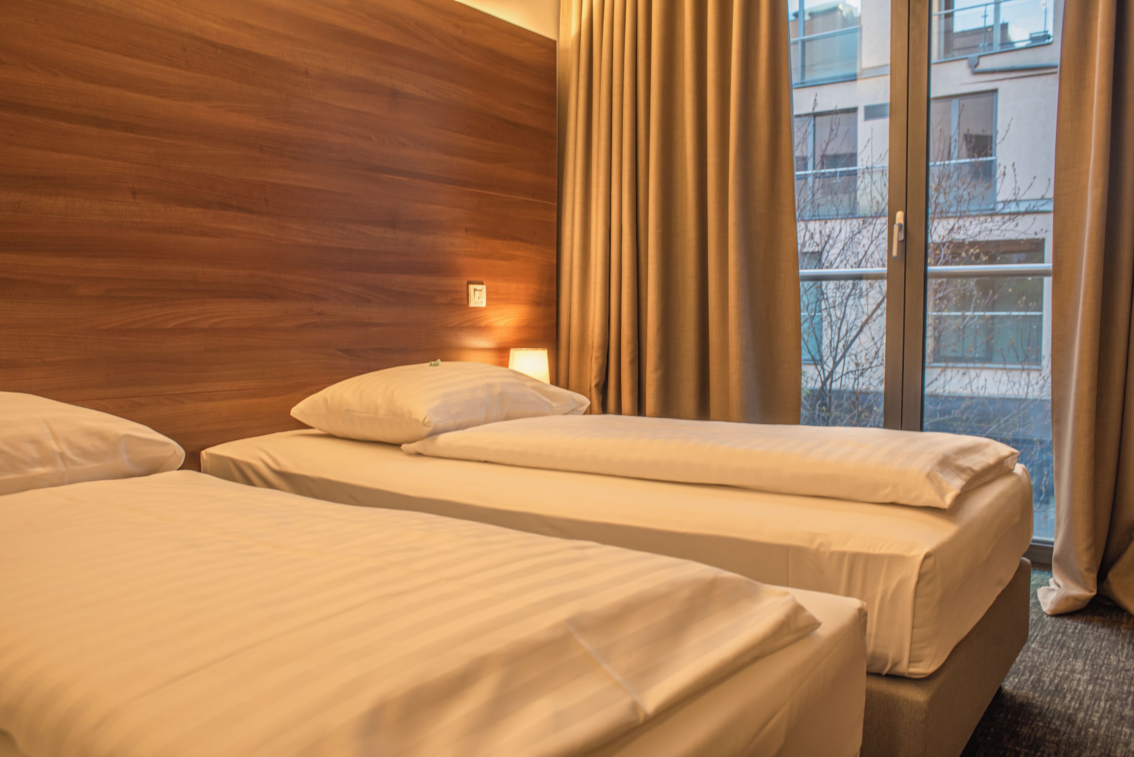 A hotel room with two beds, wooden headboard, large window with curtains, and a view of the outside.