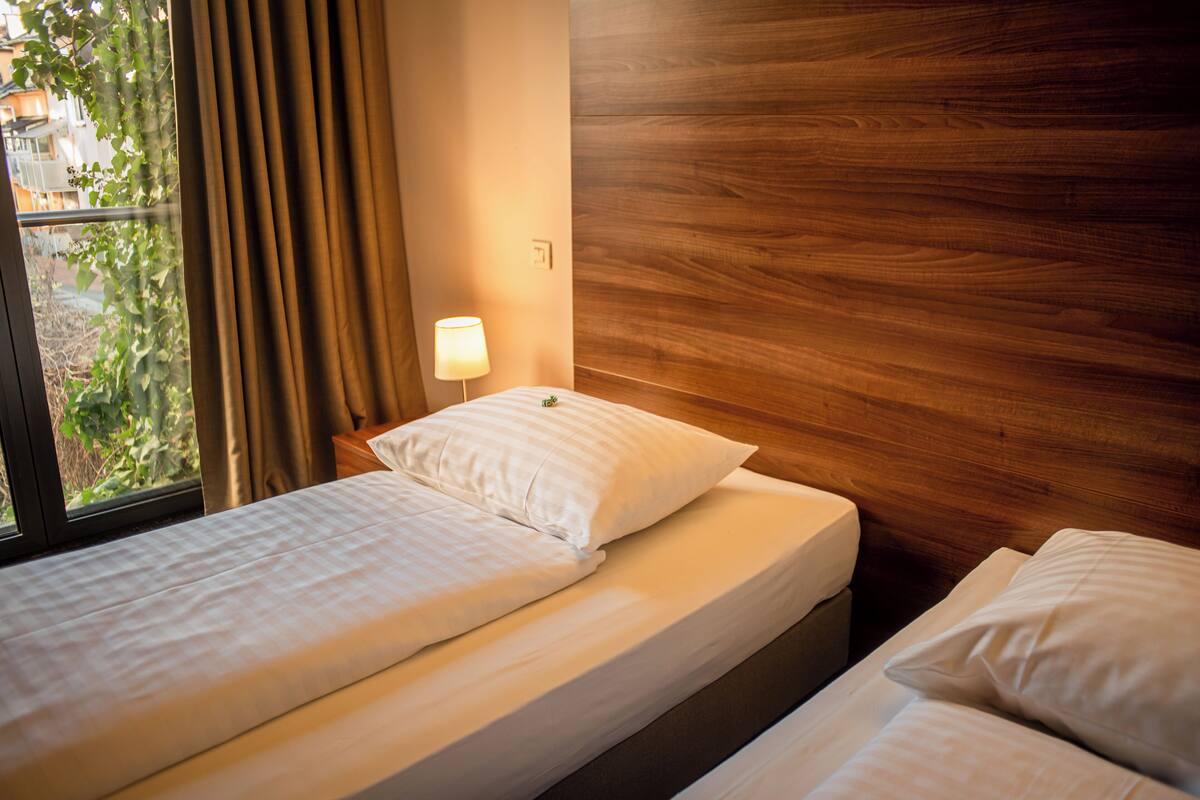 A hotel room with a wooden headboard, a window with curtains, a bedside lamp, and a bed with white bedding.