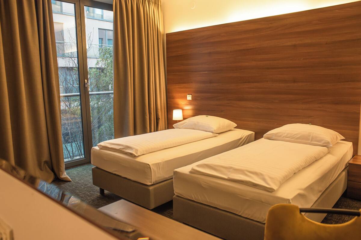 A hotel room with two single beds, a wooden headboard, a window with curtains, and a small table.