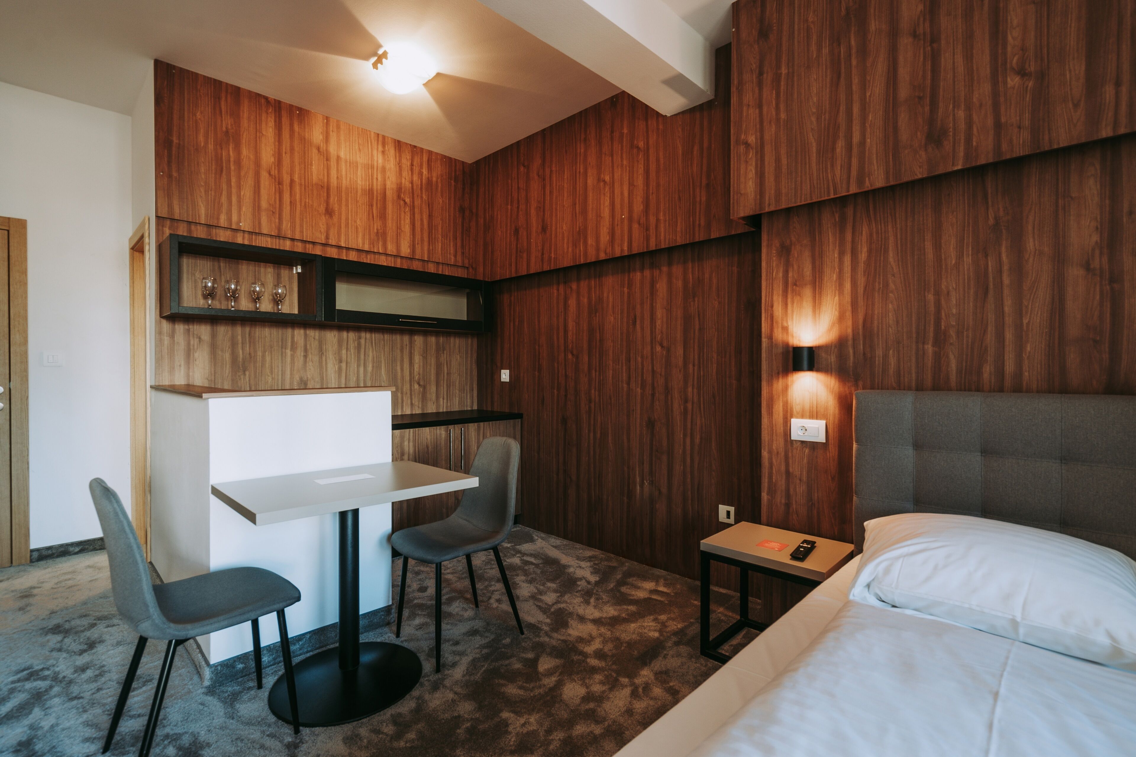 Een compacte hotelkamer met een bed, een klein bureau, twee stoelen en een houten accentmuur.