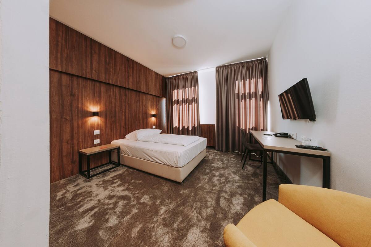A hotel room with a bed, a desk, a chair, a TV, and a small table.