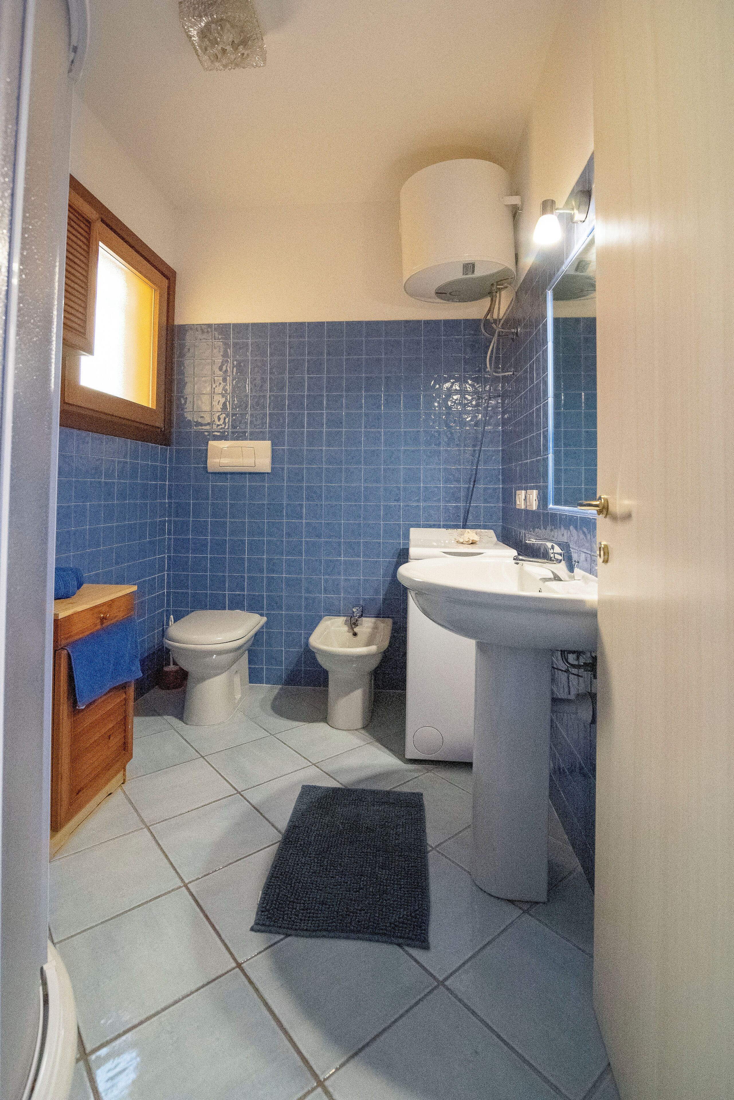 Une salle de bain comprenant des toilettes, un bidet et un lavabo, carrelée de bleu et de blanc, avec un meuble-lavabo en bois et un tapis noir sur le sol.