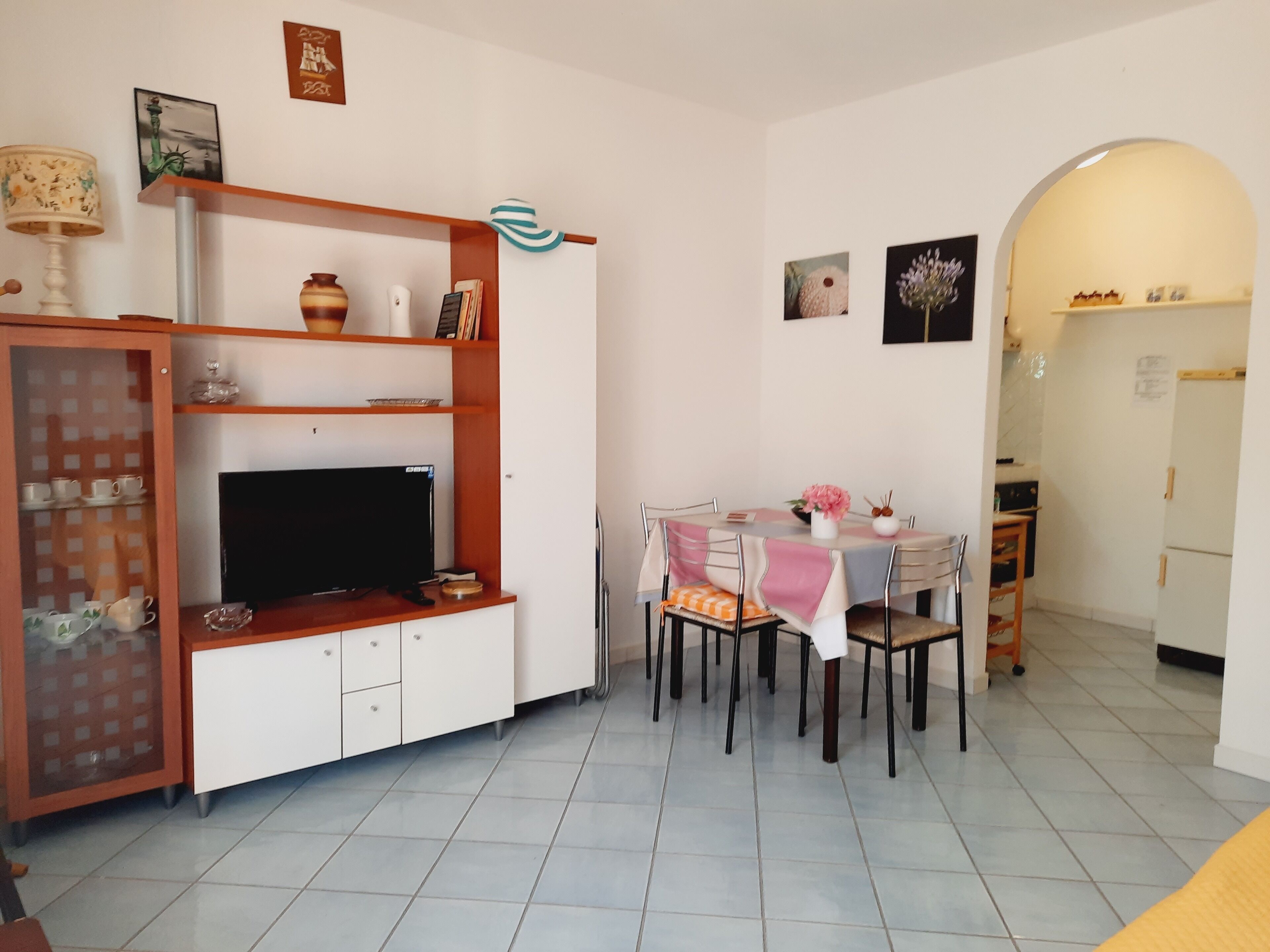 A dining area with a table set for four, a television on a cabinet, and a kitchen visible through an archway.