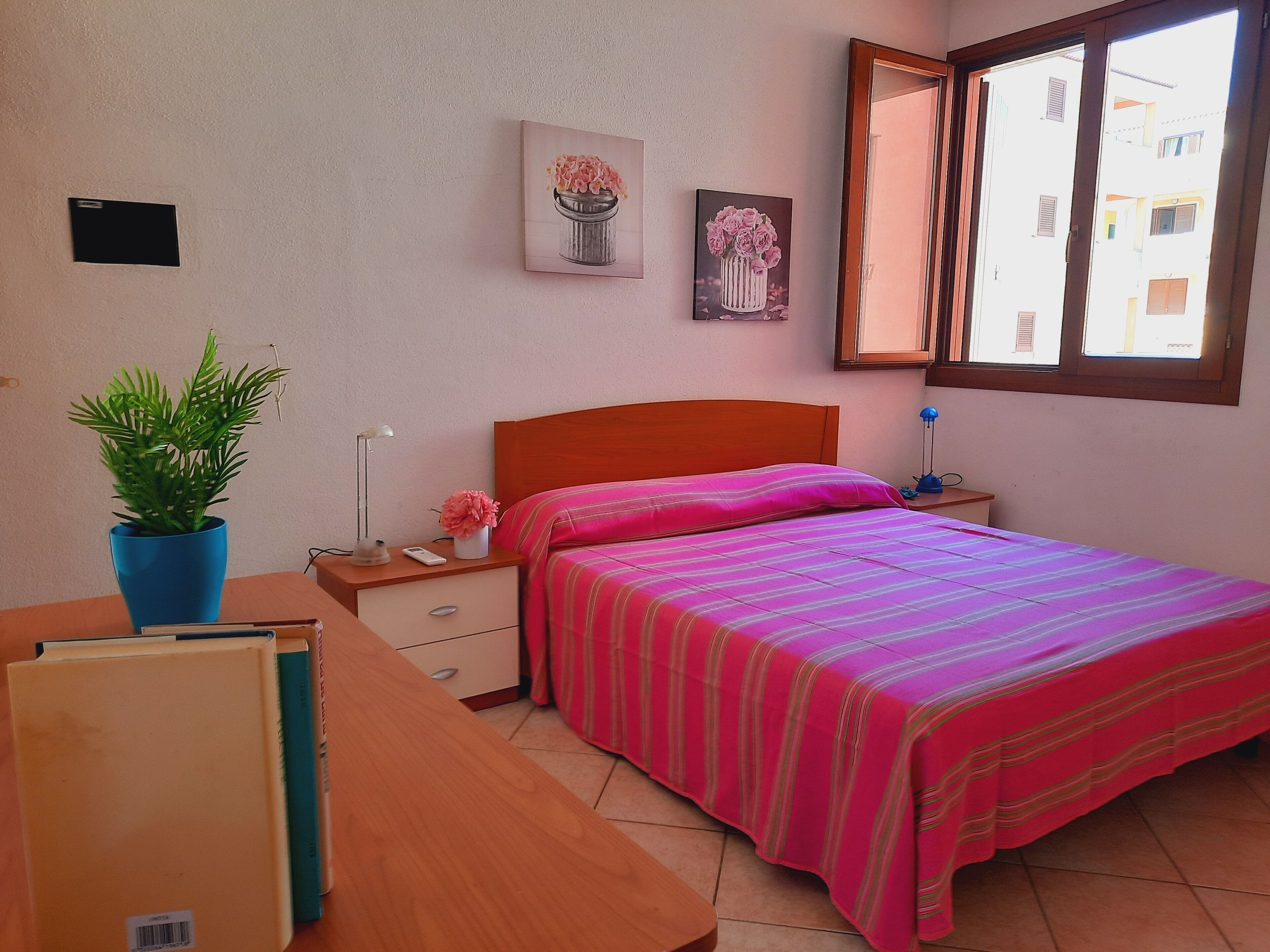 A bedroom with a bed, a nightstand, a desk, a potted plant, and two wall-mounted pictures.