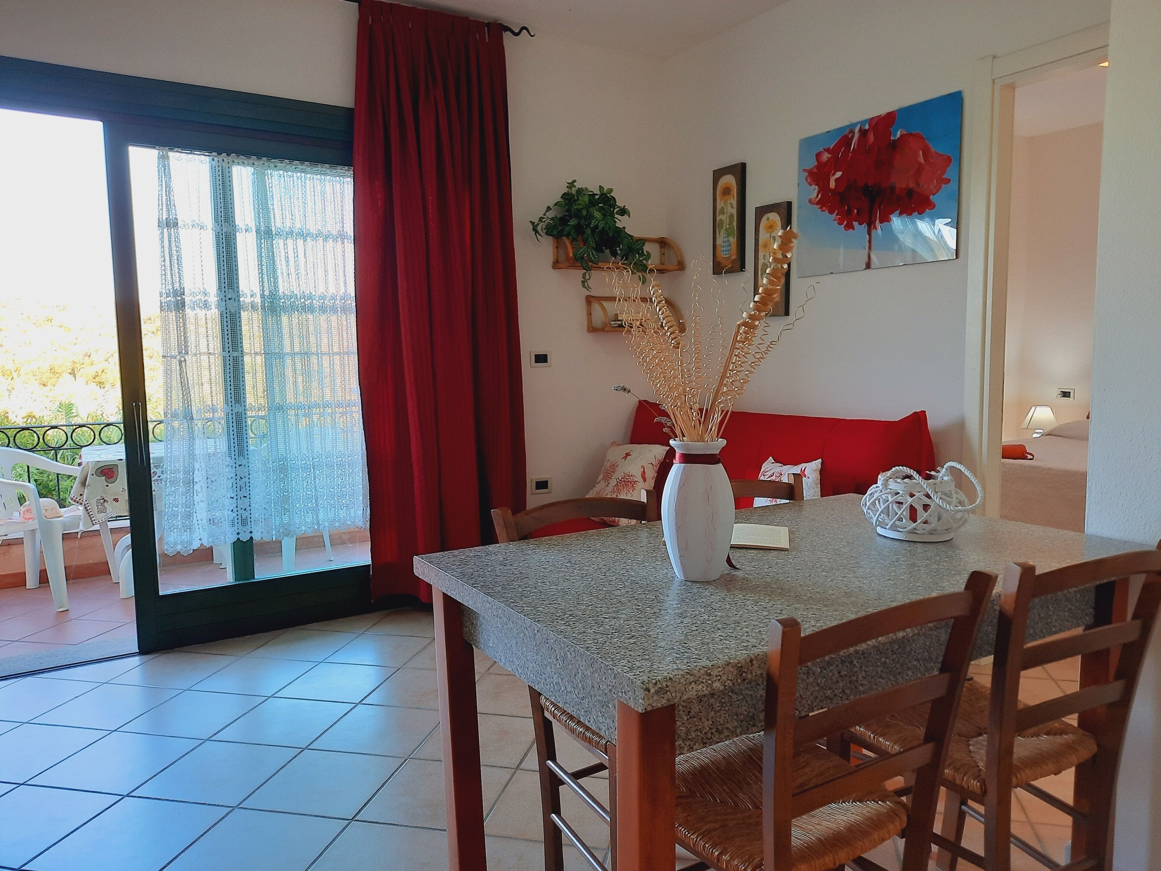 A dining area with a table, chairs, a vase with dried plants, a red sofa, and a painting on the wall.