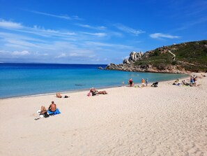 Ein Strand mit klarem, blauem Wasser, sandigem Ufer und felsigen Klippen im Hintergrund.