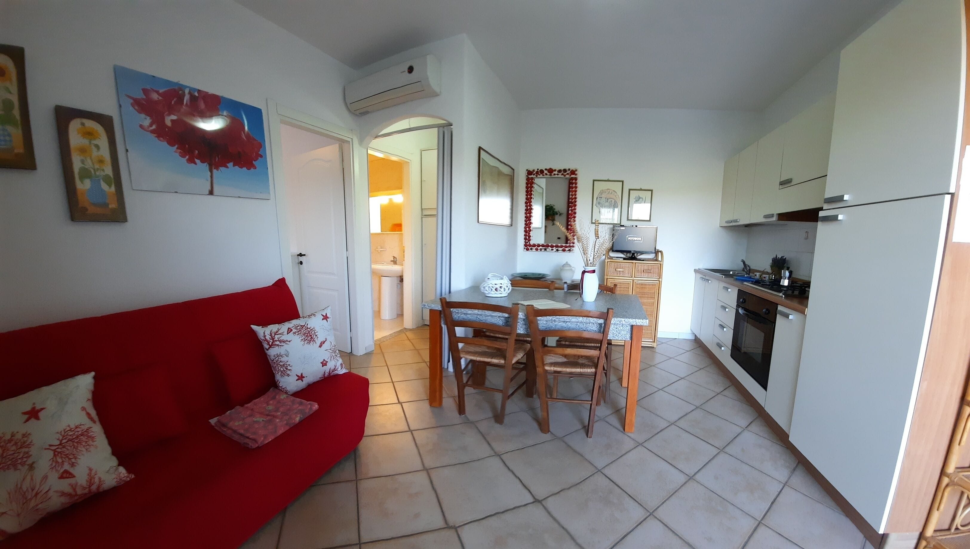Un espacio habitable compacto con un sofá rojo, una mesa de comedor con sillas, una zona de cocina con armarios y electrodomésticos, y un pasillo que lleva a un baño.
