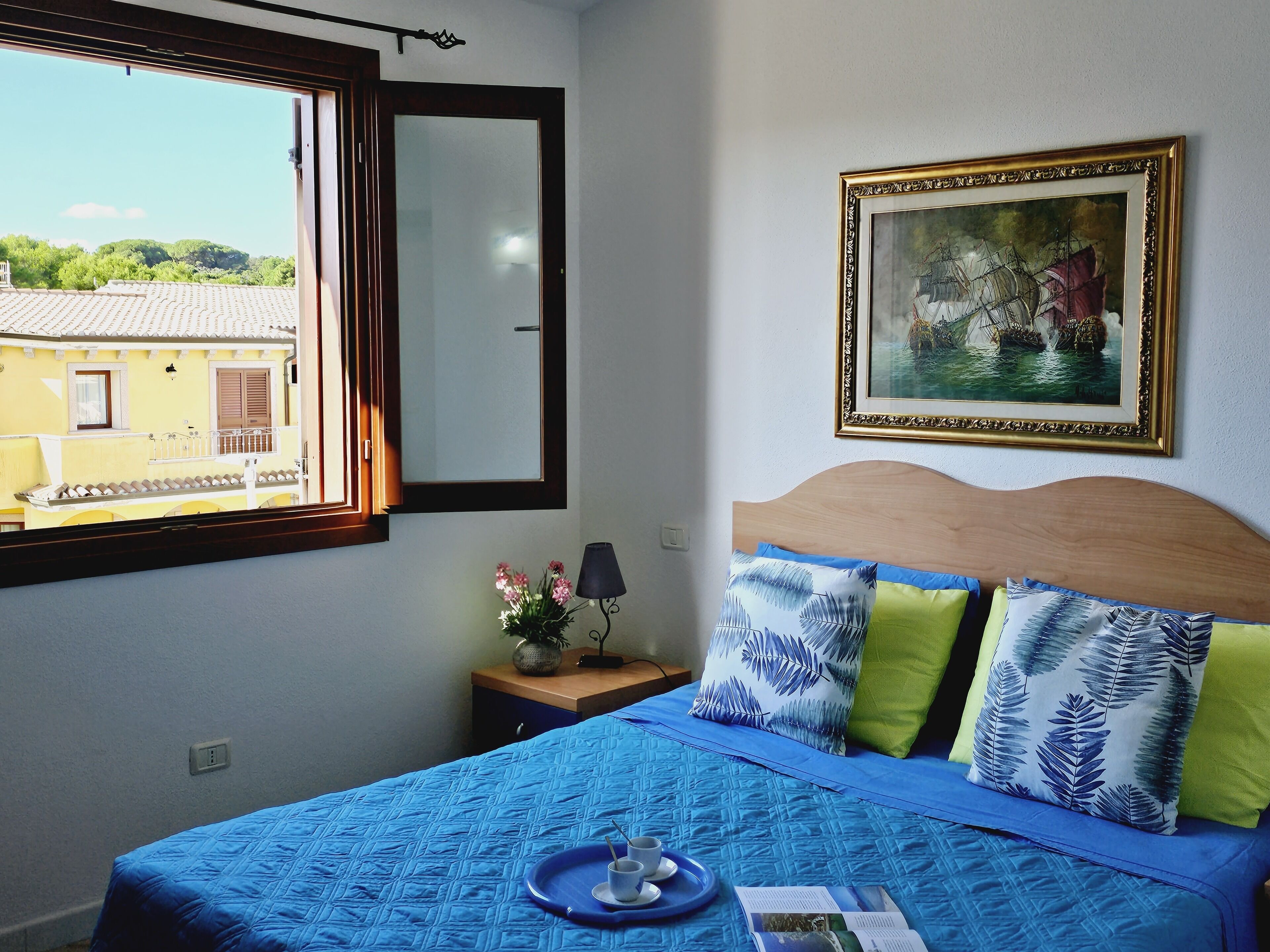 Een slaapkamer met een bed, een raam, een nachtkastje, een lamp, een lijst met een schilderij en uitzicht op gebouwen.