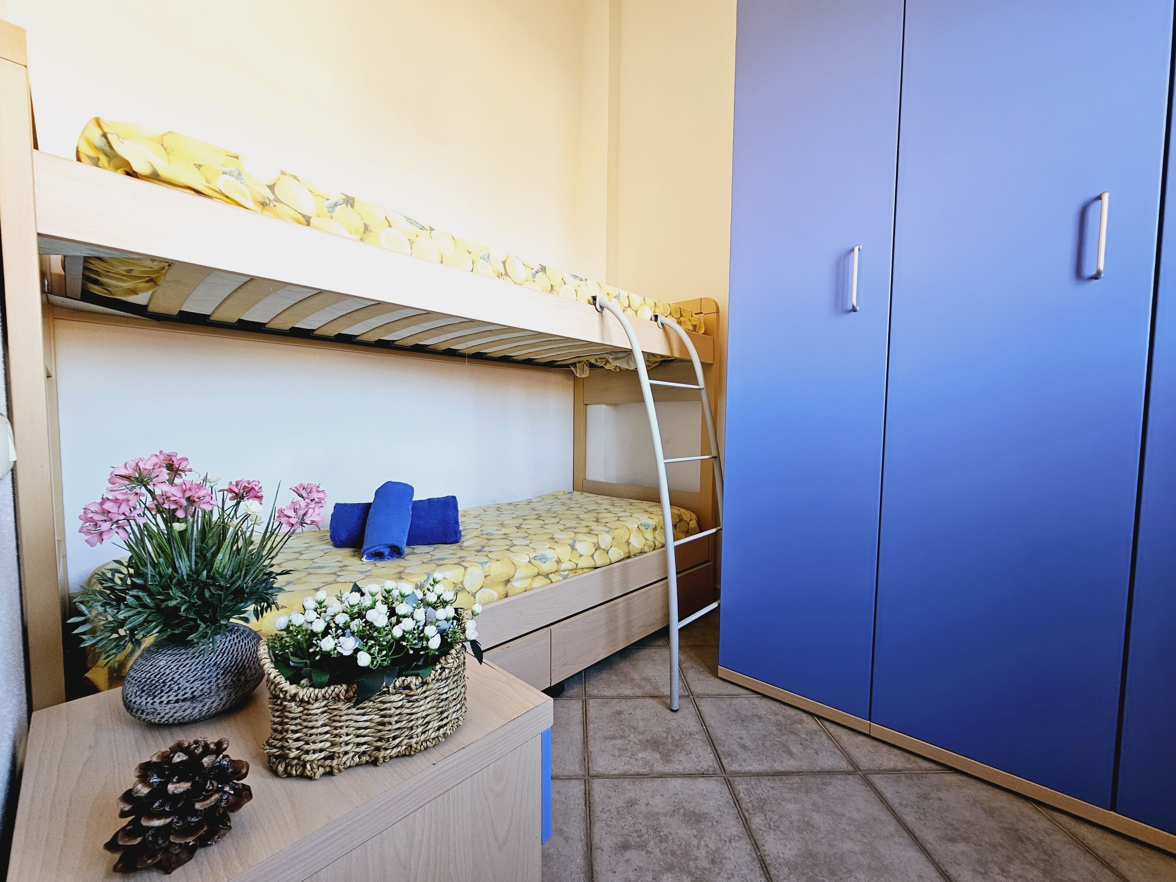Una cama litera con escalera, un armario azul y un escritorio de madera con una piña y flores.