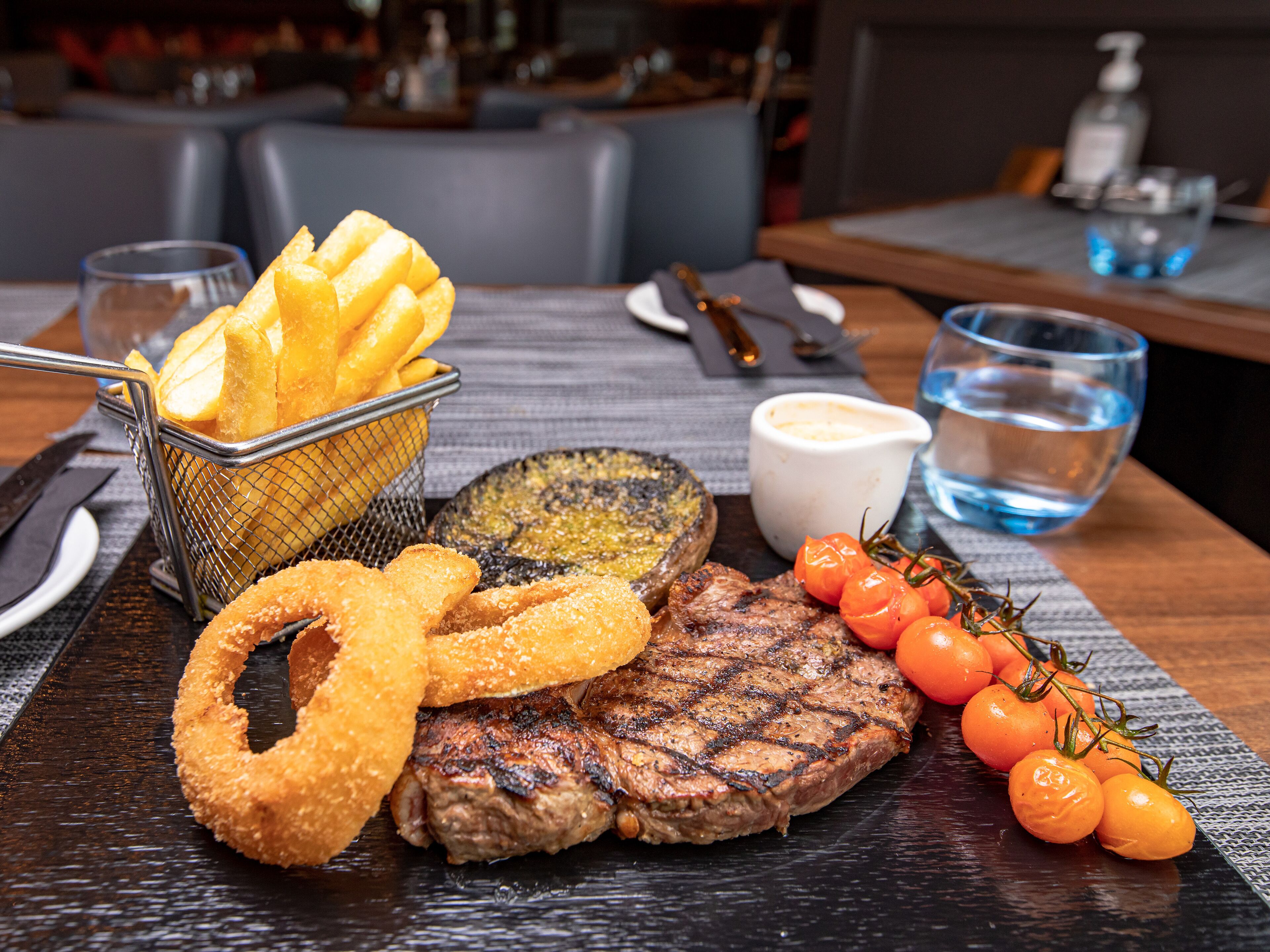 A meal consisting of grilled steak, onion rings, fried potatoes, and cherry tomatoes on the side.