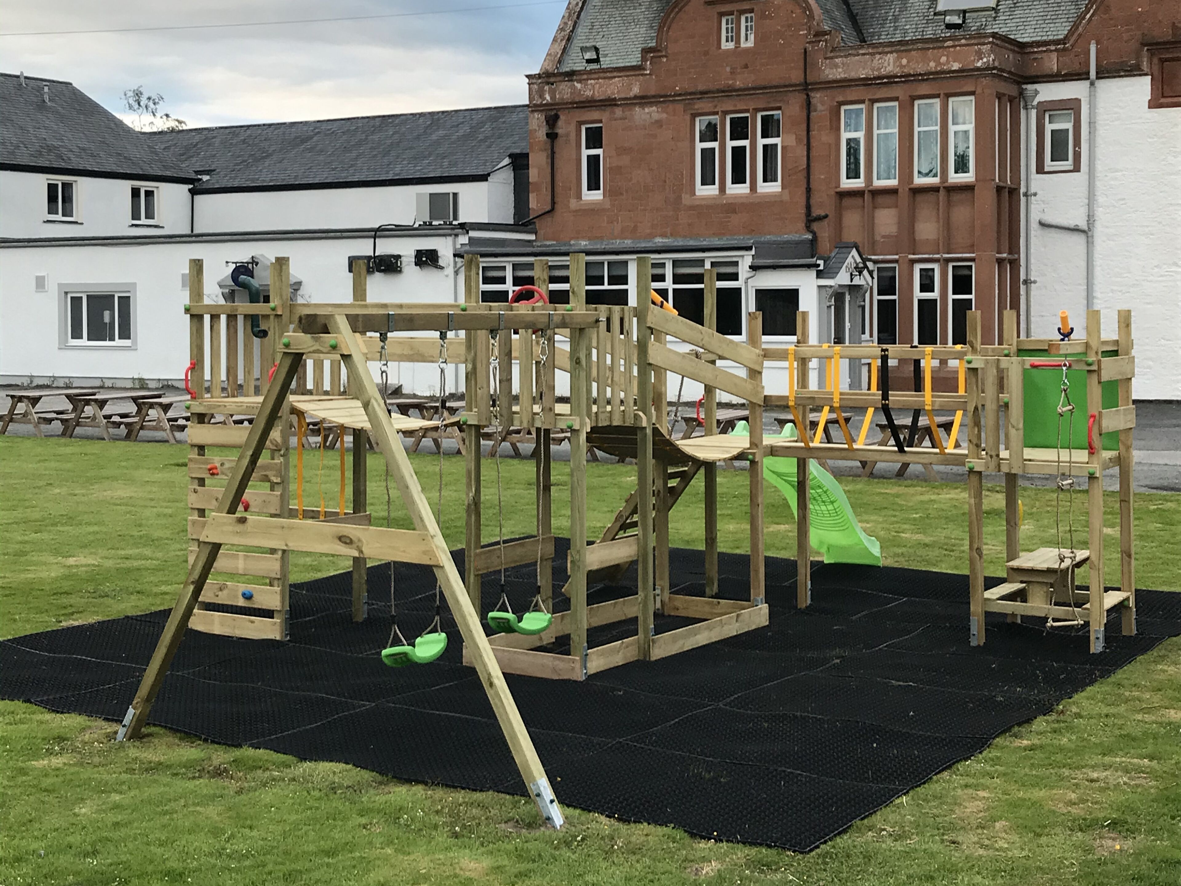 Un terrain de jeux équipé de structures d’escalade et d’un toboggan, installé sur un tapis noir devant un bâtiment doté de nombreuses fenêtres et d’une façade en briques.