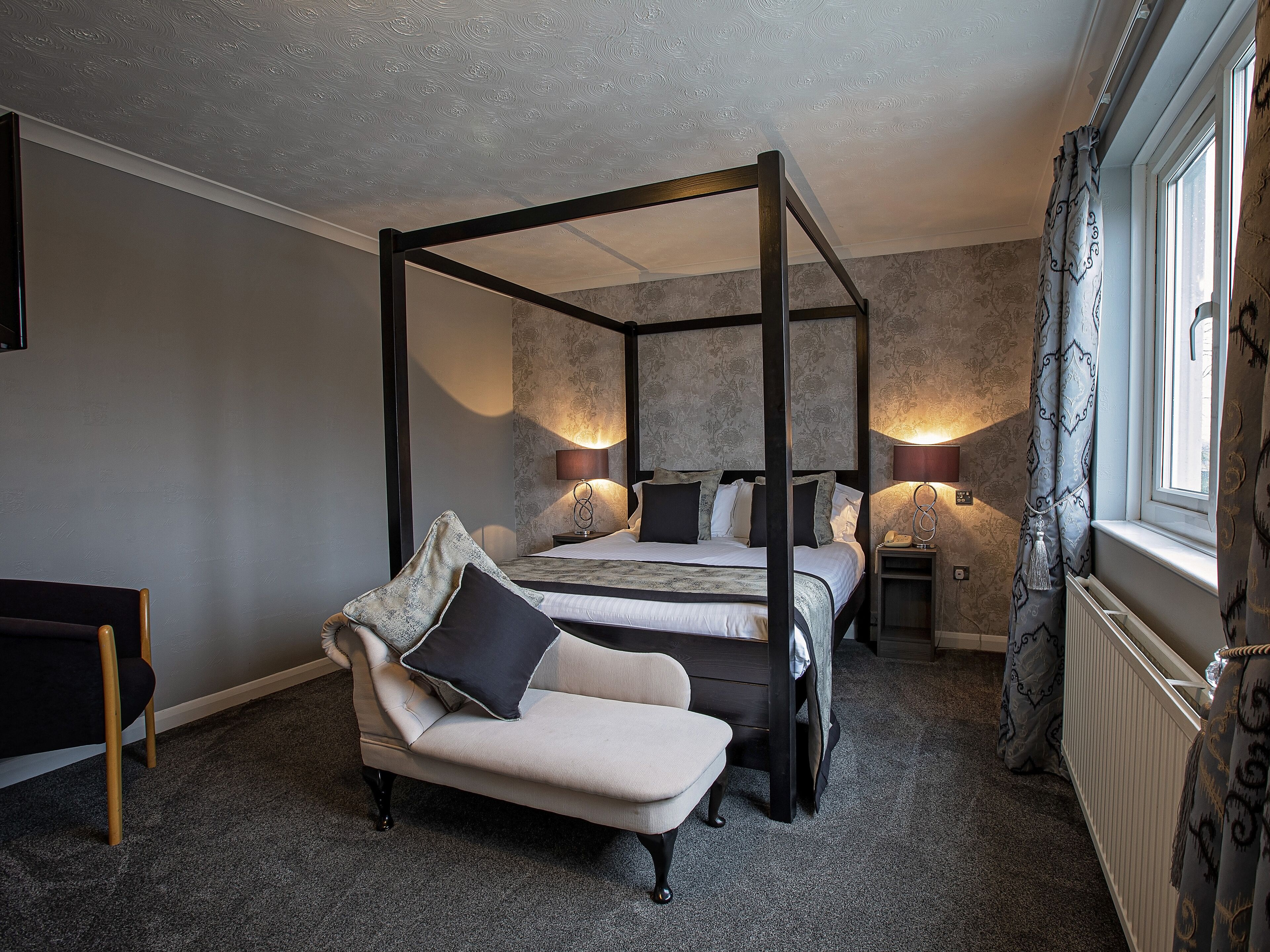 A four-poster bed with a canopy, two bedside tables with lamps, a chaise lounge, and a window with patterned curtains.