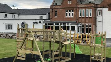 Guests can enjoy a playground with a swing set and climbing frame, set amidst a traditional building with a pitched roof.