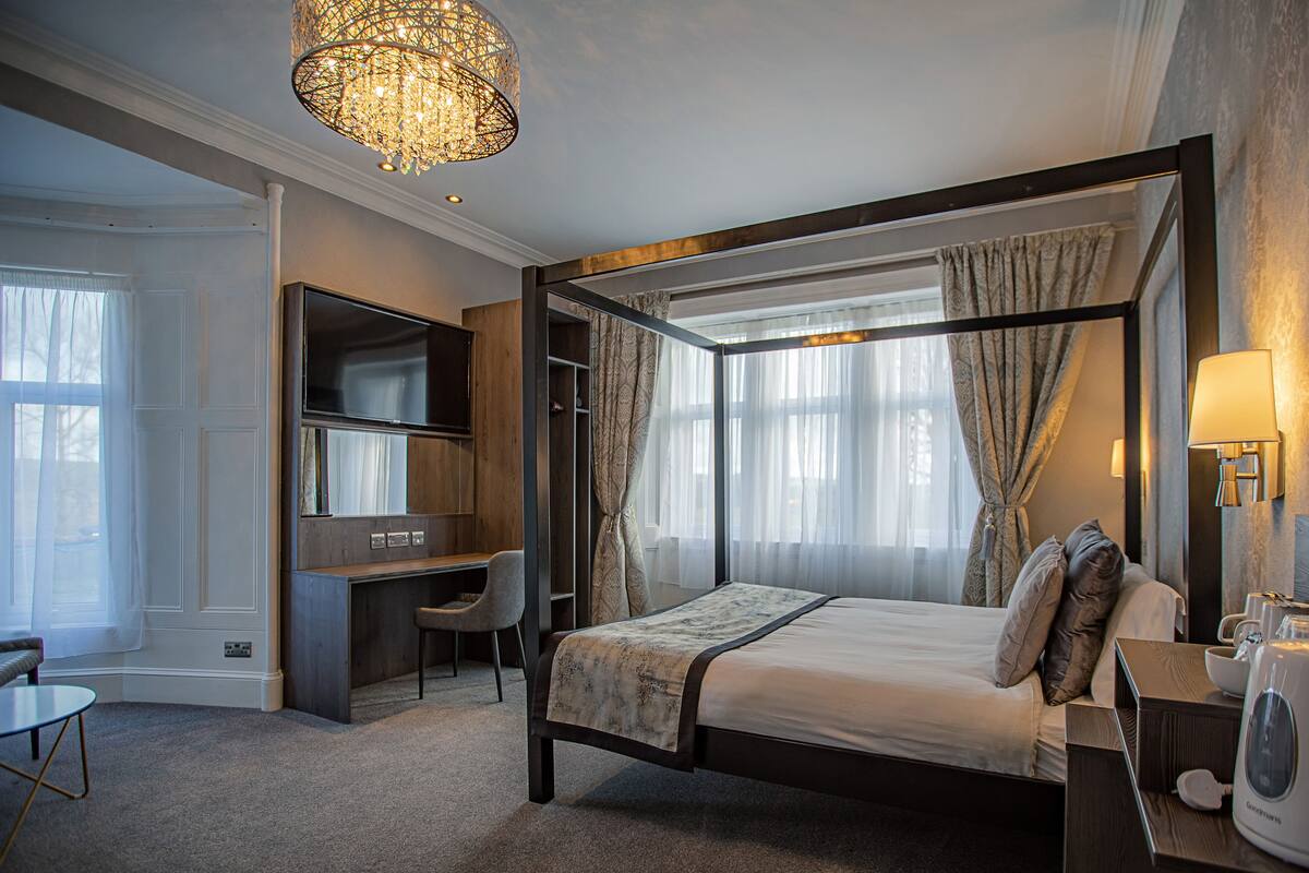 A bedroom with a four-poster bed, a desk, a chair, a television, and a chandelier.