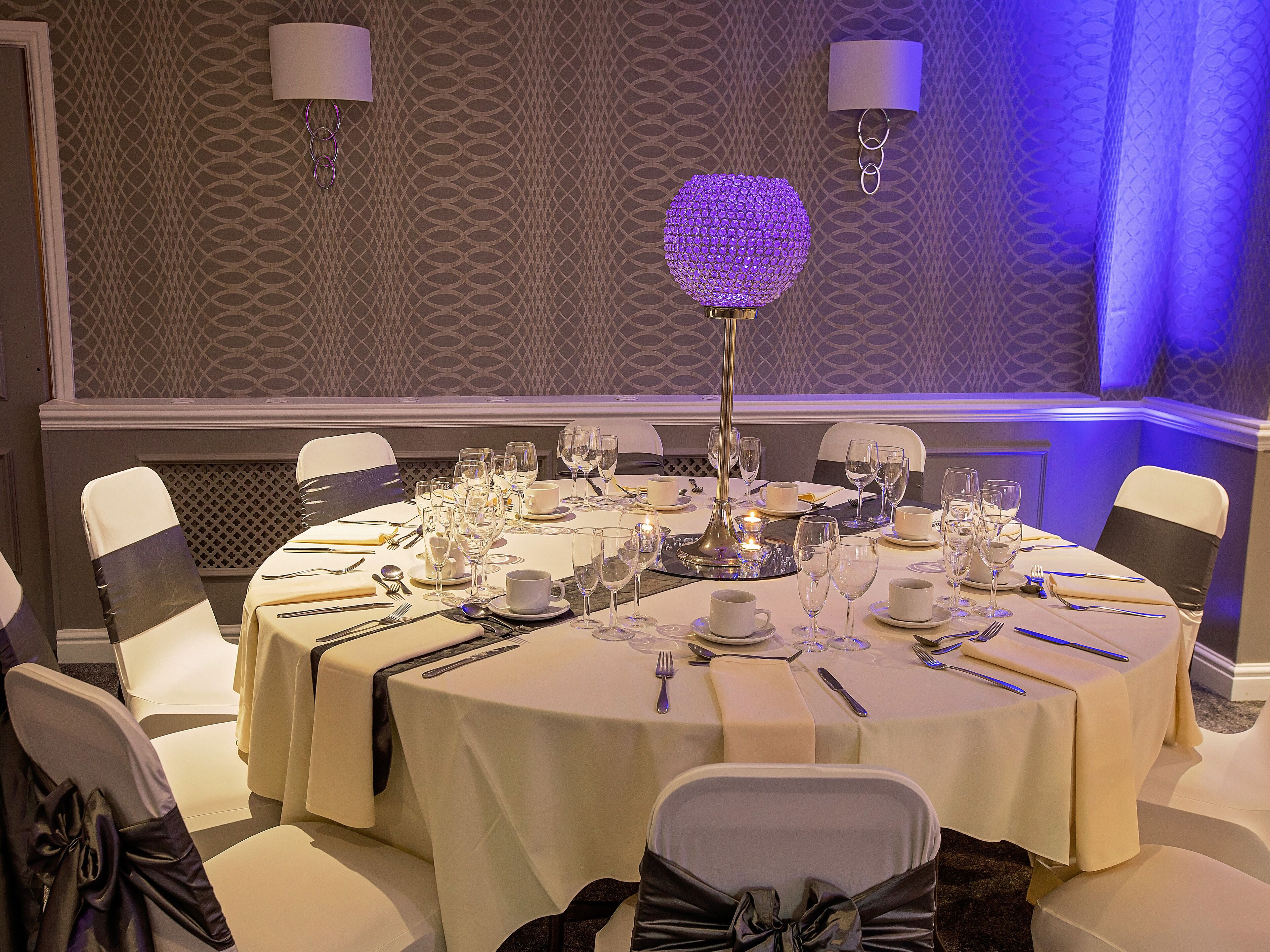A formal dining room with round tables, white tablecloths, and chairs with black sashes.