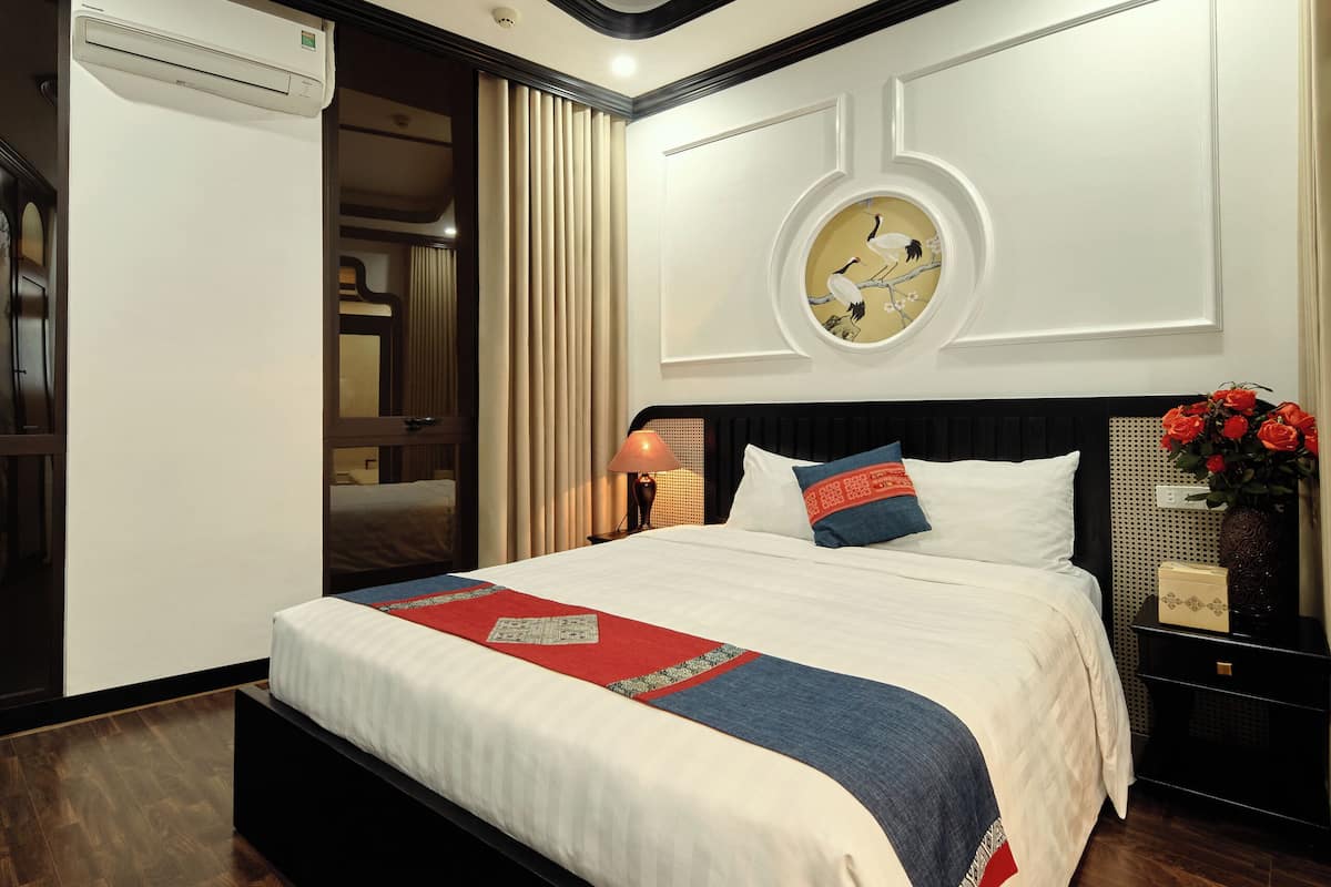 Una habitación de hotel moderna con una cama grande, un aire acondicionado de pared, un adorno en la pared y una mesita de noche con un florero.