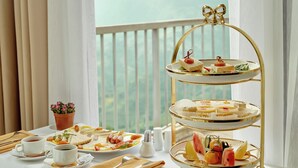 A three-tiered dessert stand on a table with a white tablecloth, a potted plant, and a view of the sea through a balcony. - Amazing Hotel Sapa (Sa Pa)