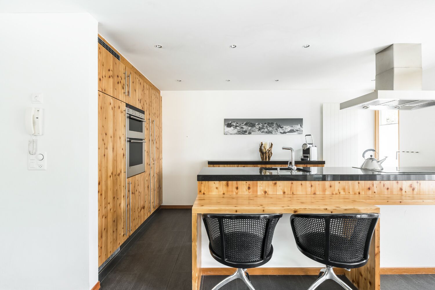 A modern kitchen with wooden cabinetry, a black countertop, and two black mesh chairs.