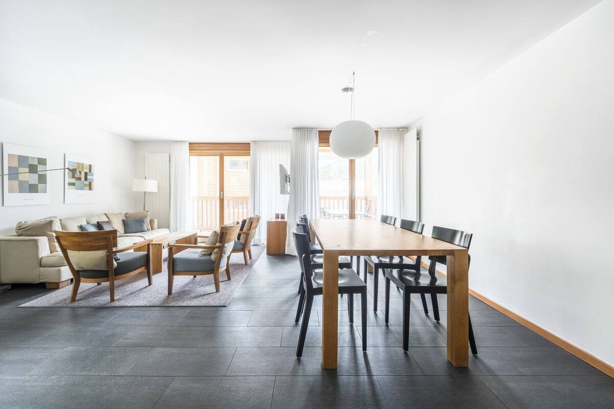 Uma sala de estar moderna com uma mesa de jantar de madeira, cadeiras e um sofá. Há uma janela grande com cortinas, uma luminária e obras de arte na parede.