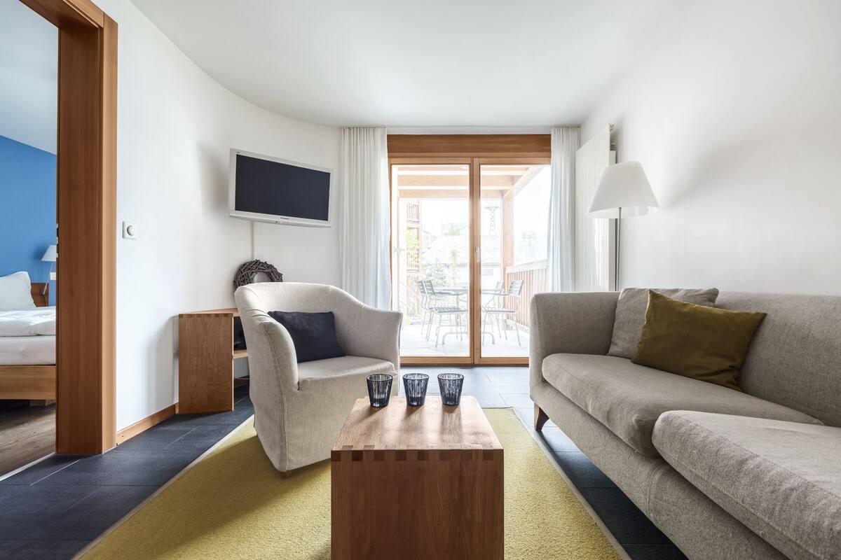 Quarto de hotel moderno com cama, sofá, poltrona, televisão e vista exterior.
