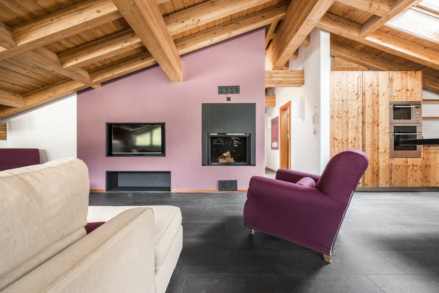 Un soggiorno moderno con soffitto alto, travi in legno, un camino, una televisione e un angolo cottura con pannelli di legno.