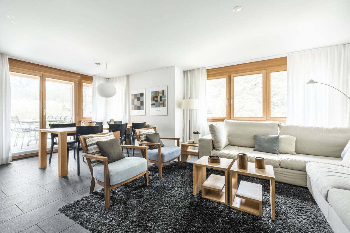 Uma sala de estar moderna com sofá, mesa de centro e cadeiras. Há uma área de jantar com mesa e cadeiras. Grandes janelas proporcionam iluminação natural.