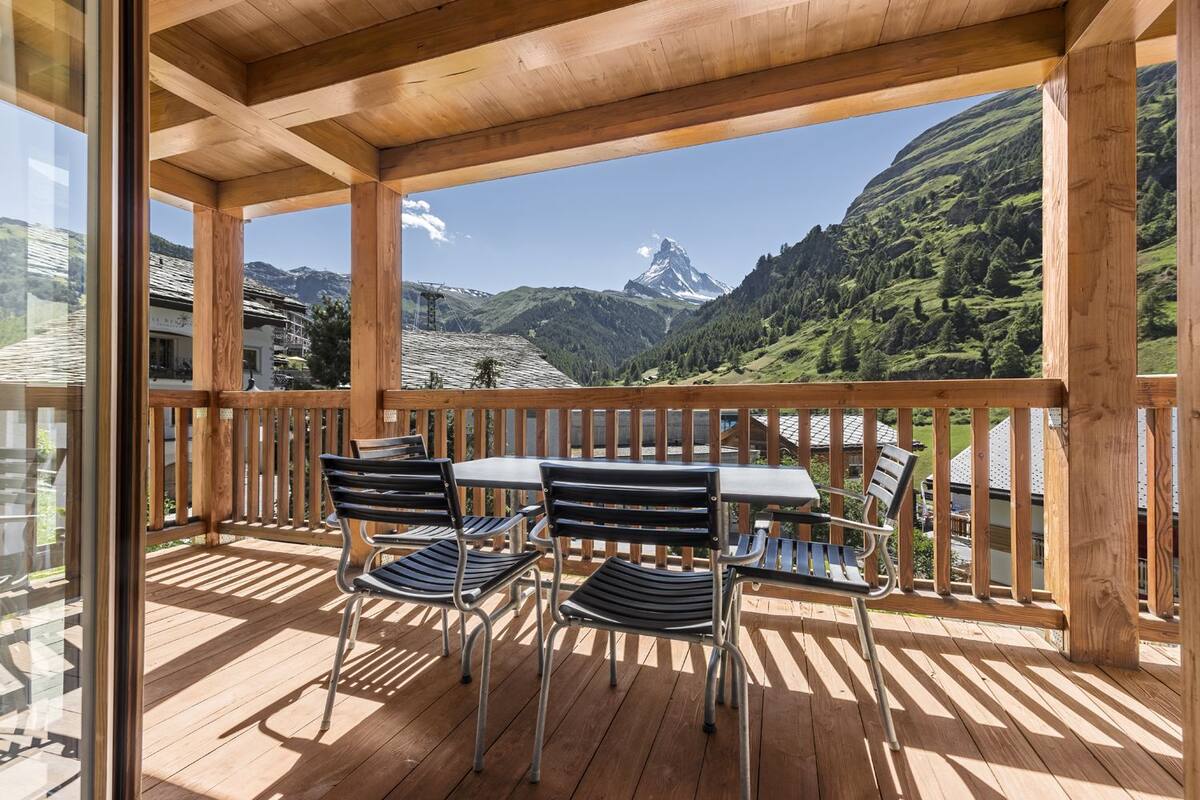 Um deck de madeira com mesa e cadeiras, com vista para uma paisagem montanhosa e um pico imponente.