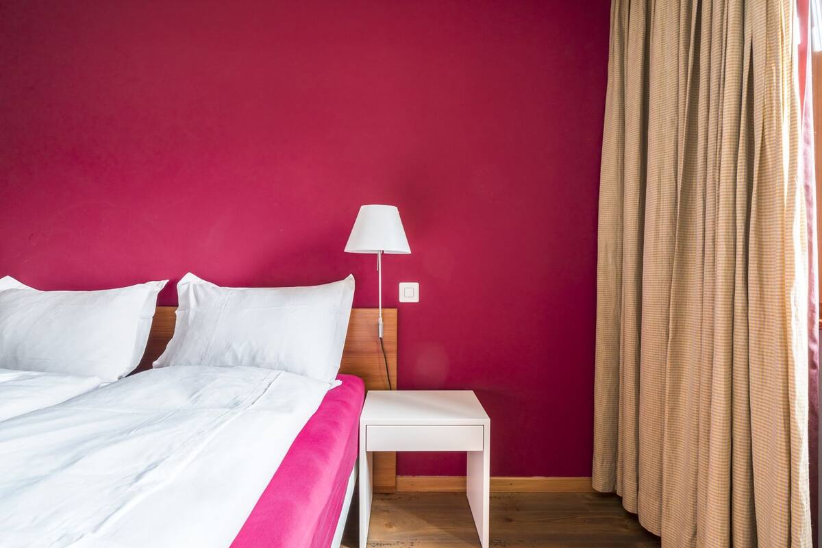 Quarto de hotel com cama, mesa de cabeceira, abajur e cortina.