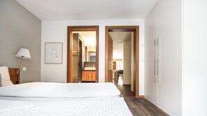 A hotel room with a large bed, a nightstand, a lamp, a framed picture on the wall, and a wooden door leading to a bathroom and a dining area. - Ari Resort (Zermatt)