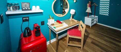 A compact room with teal walls, a wooden floor, a small table with a red chair, a red appliance, a mirror, and a shelf with various items.