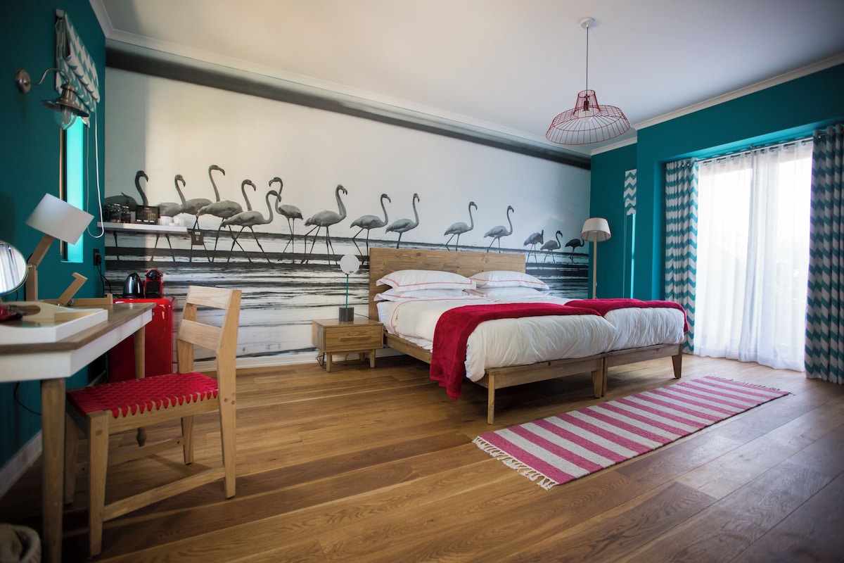 A bedroom with a bed, a desk, a chair, a nightstand, and a wall mural of flamingos.