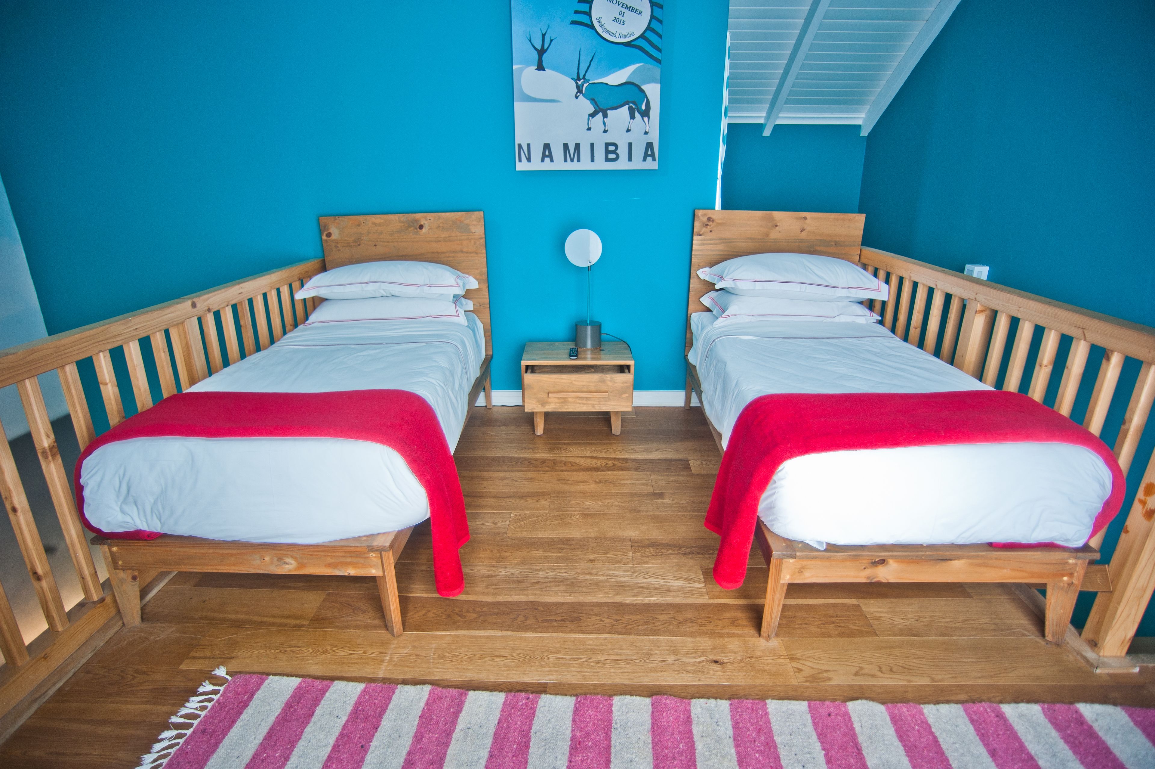 A room with two wooden beds, a small wooden nightstand, a wall poster of Namibia, and a striped rug on the floor.