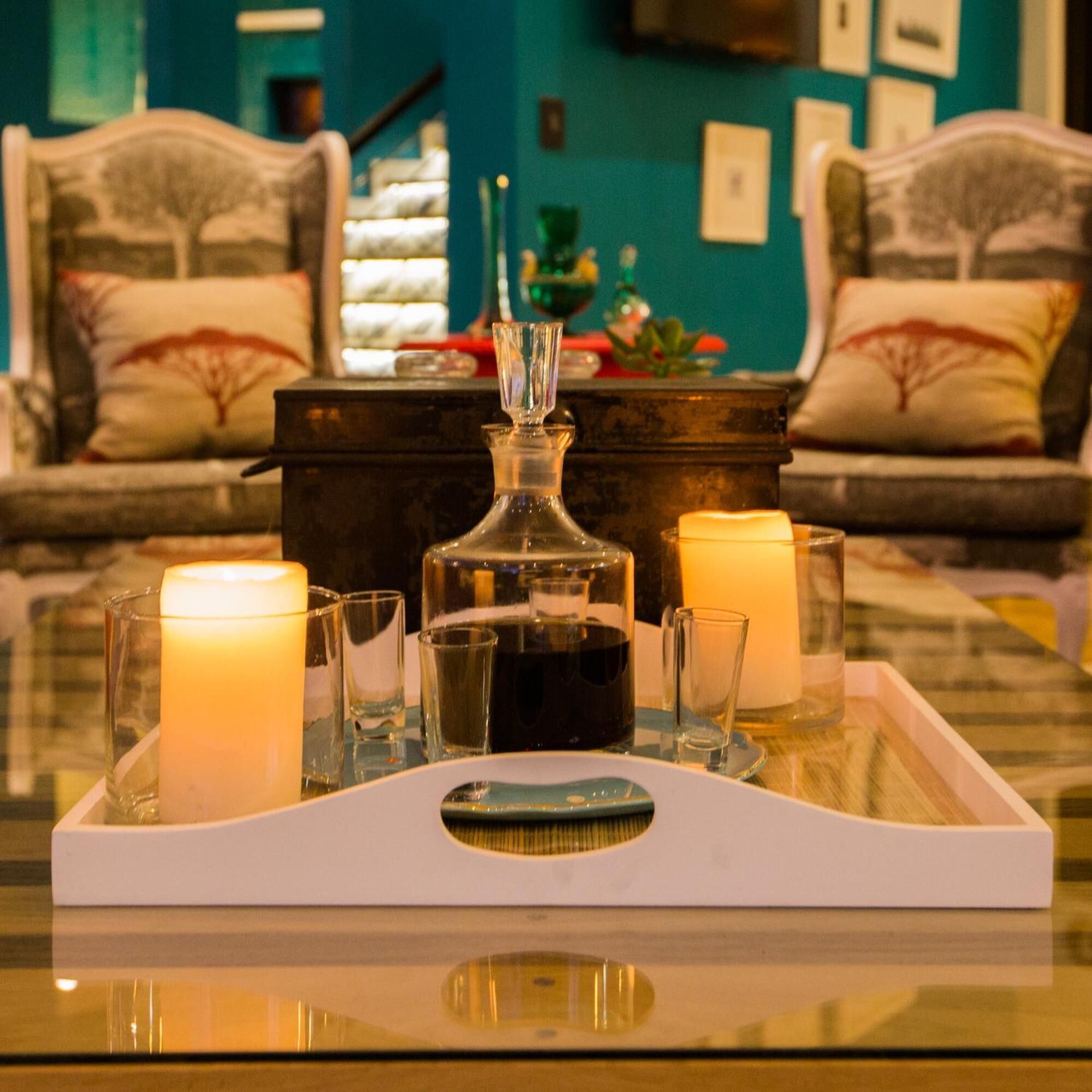 Un plateau avec une carafe, des bougies et des verres est posé sur une table dans une pièce où se trouvent des canapés à motifs et un mur de couleur turquoise.