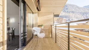 A wooden balcony with a glass door leading to a dining area, a white chair, and a view of a snowy mountain and buildings.