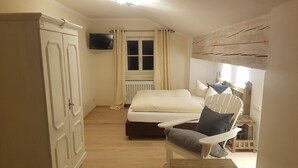Une chambre à coucher comprenant un lit, une chaise, une petite table, une armoire, un téléviseur et une fenêtre avec des rideaux.