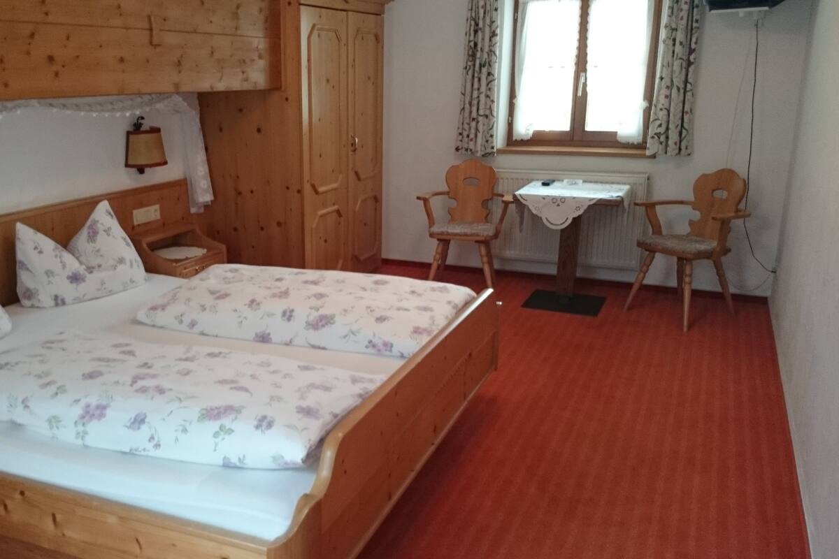 Un dormitorio con cama de madera, dos sillas, una mesita y una ventana con cortinas.
