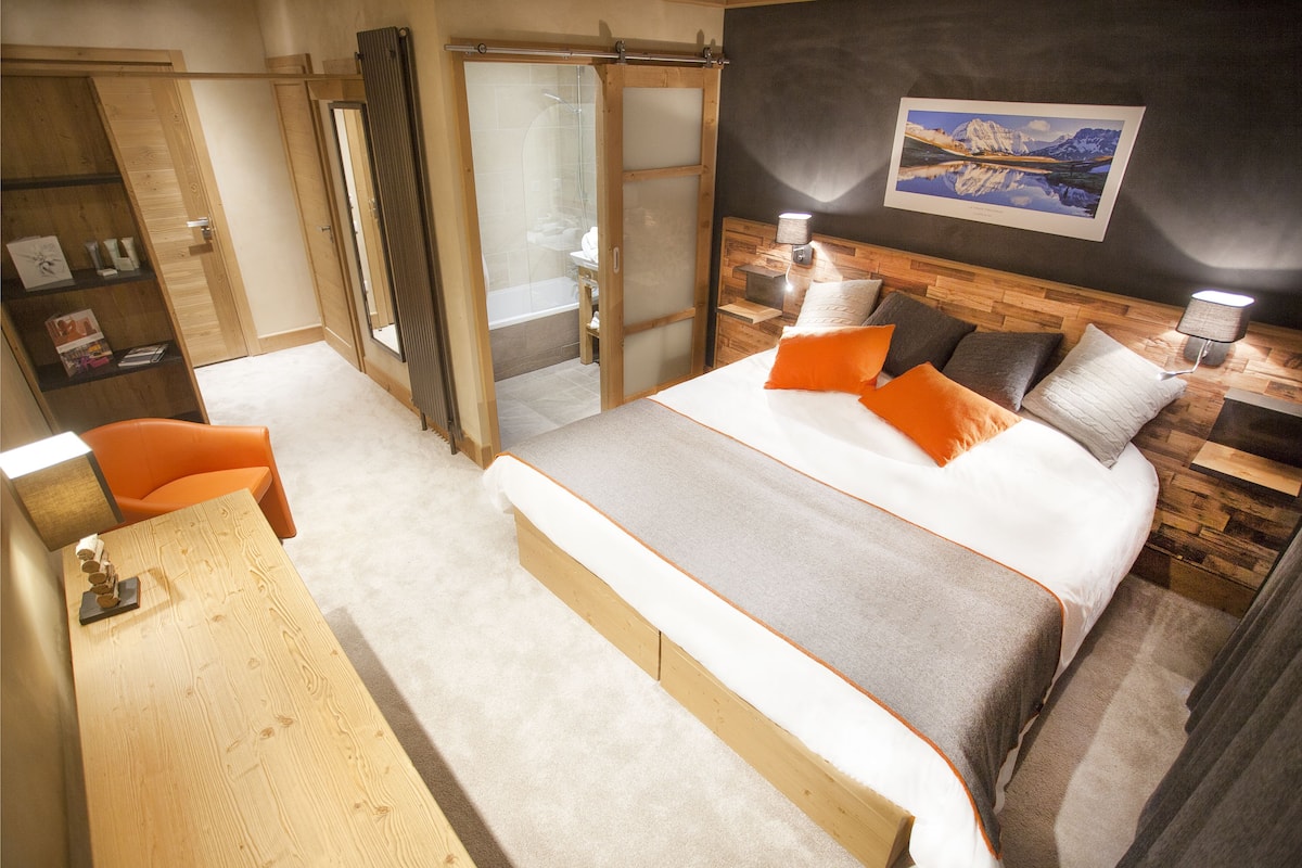 Quarto com cama grande, escrivaninha de madeira, cadeira, guarda-roupa e banheiro com chuveiro de vidro.