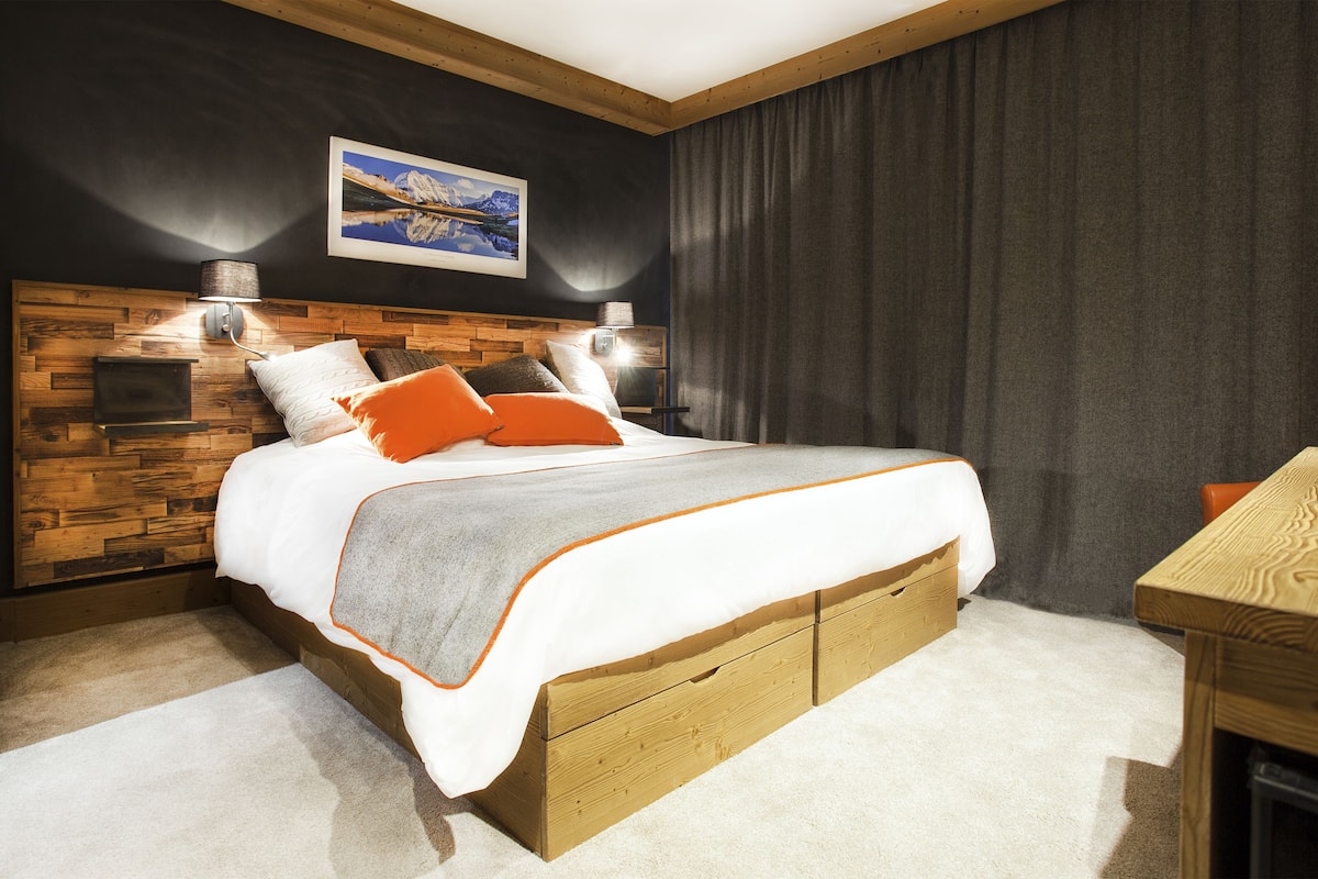 Quarto moderno com cabeceira de madeira, uma cama grande com roupa de cama laranja e branca, duas luminárias de mesa e uma TV na parede.