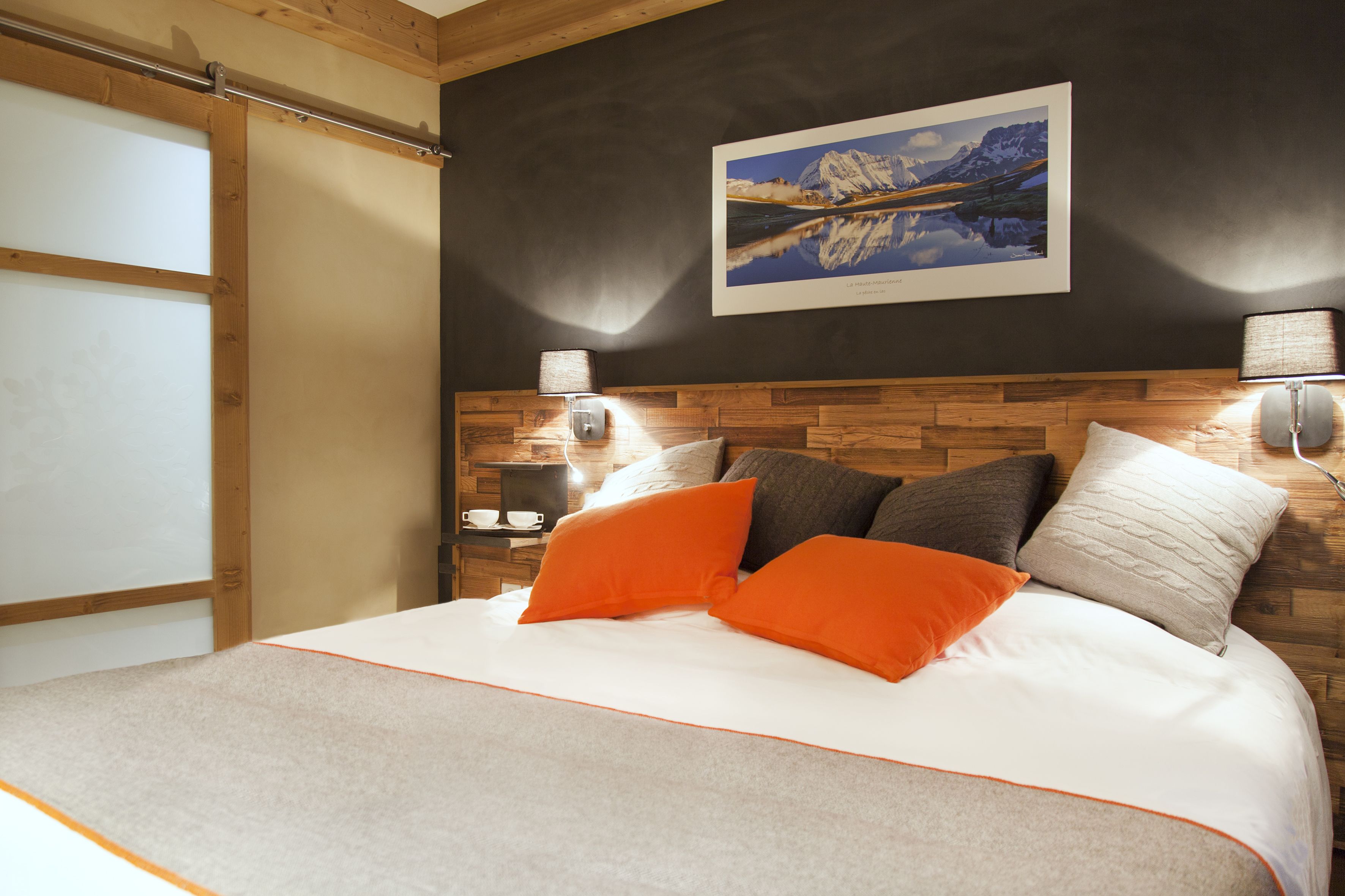 A bedroom with a wooden headboard, a large bed with white bedding and orange pillows, a framed picture on the wall, and two bedside lamps.
