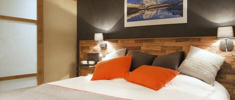 Een slaapkamer met een houten hoofdeinde, een groot bed met wit beddengoed en oranje kussens, een ingelijst kunstwerk aan de muur en twee bedlampen.