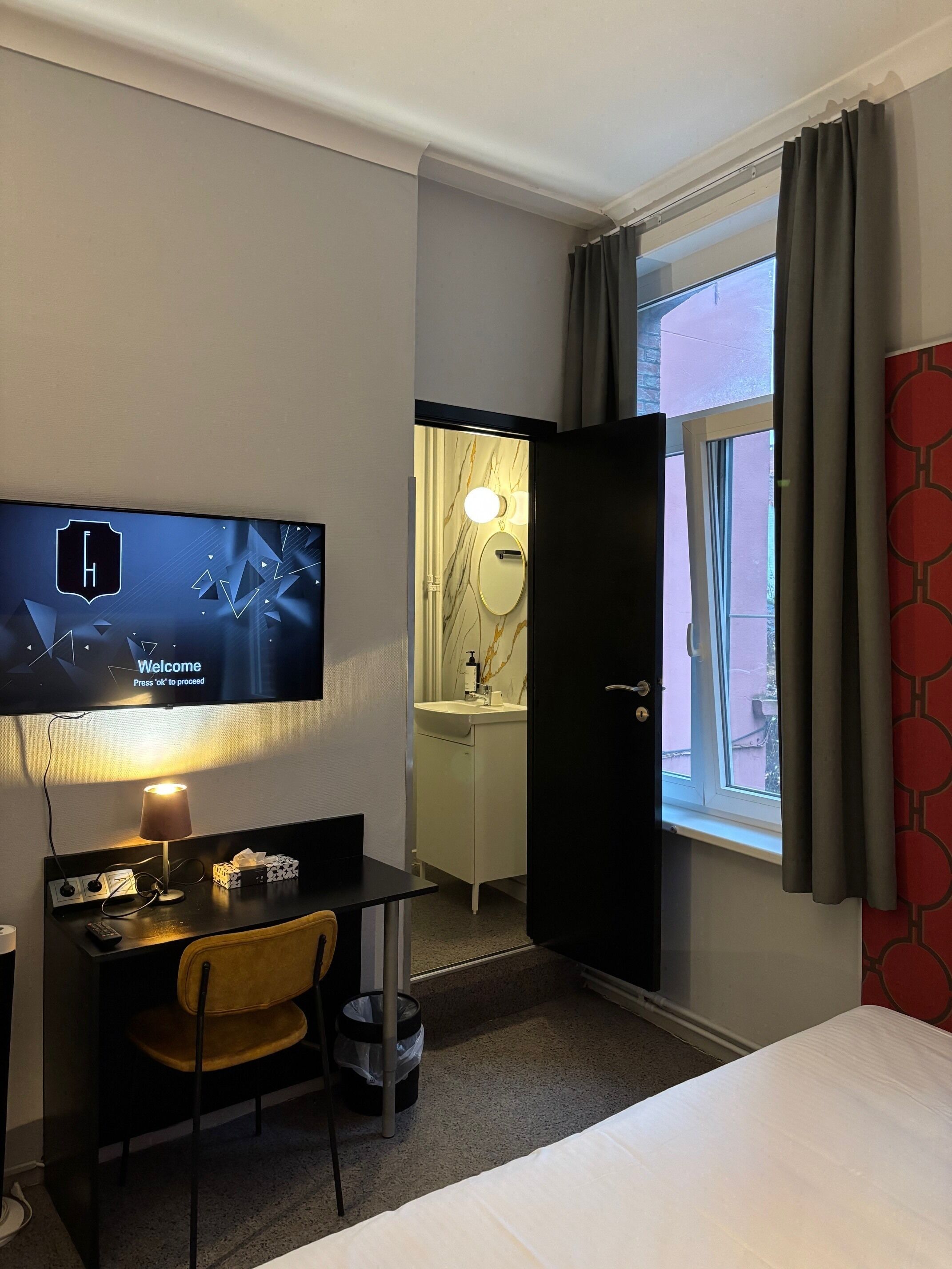 Une chambre d’hôtel comprenant un lit, un bureau avec une chaise, une télévision, une table de chevet, une lampe, une poubelle et une fenêtre avec des rideaux.