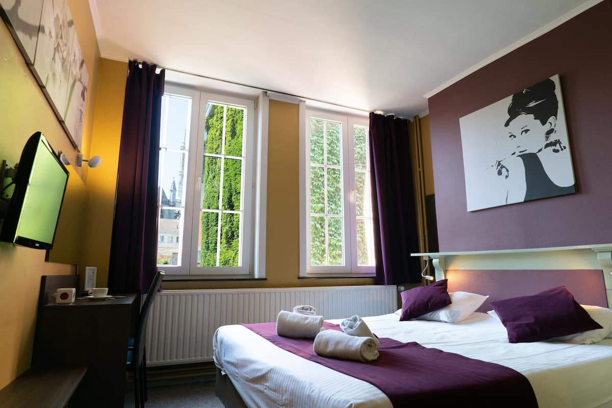 Habitación de hotel con cama, escritorio, silla, televisor y ventana con cortinas.